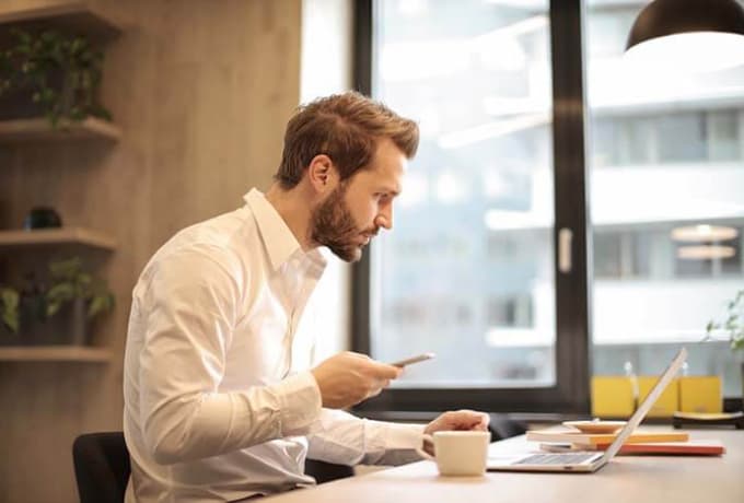 man is sitting at the table holding phone in right hand and looking at the screen of the laptop